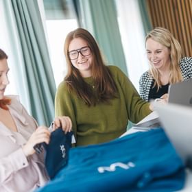 Three women stand at a table with blue clothing items laid out and look at them together. One woman holds a piece of clothing while the others smile and look at it; laptops are visible in the background.