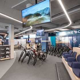 The image shows the bike rental area inside the Bründl Sports AreitXpress valley station shop with several e-bikes, bike helmets, seating, and a service area. The texts “#BRÜNDLFAN,” “RETURN,” and “ONLINE BOOTS,” as well as Bründl Sports logos, are visible in the image.