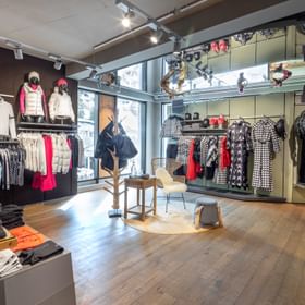 Das Bild zeigt eine stilvolle Verkaufsfläche in einem Bründl Sports Shop, wo Wintermode wie Jacken, Pullover und Accessoires präsentiert werden. Schaufensterpuppen tragen farbenfrohe Outfits, und in der Mitte steht ein dekoratives Sitzmöbel mit einem kleinen Tisch, was eine gemütliche Atmosphäre schafft.