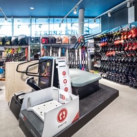 Innenansicht des Bründl Sports Shops Planet Planai in Schladming mit einer großen Auswahl an Skischuhen, Ski und Skistöcken. Im Vordergrund steht ein BootDoc Fußanalysegerät für die Skischuhanpassung, daneben befindet sich eine gepolsterte Sitzbank. Große Fenster und eine blaue Decke prägen den Verkaufsbereich.