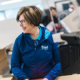 A woman wearing glasses and a blue jacket with the text “Bründl Sports” stands at a worktable. She looks to the side smiling, with boxes and work materials visible in the background.