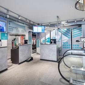 Zu sehen ist der Skiverleihbereich im Shop Bründl Sports Saalbach life.style mit mehreren Service- und Verleihstationen, Ski, Skistöcken und Bildschirmen. Sichtbar sind unter anderem die Texte „#BRÜNDLFAN“, „RENT SKI“, „PREISE Ski & Stöcke“, „ONLINE TICKET“ und „WIR BEWEGEN MENSCHEN“; rechts befindet sich eine Rolltreppe.