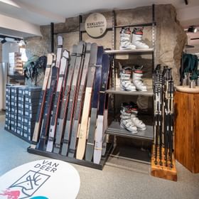 Das Bild zeigt eine Skiausrüstungsausstellung in einem Bründl Sports Shop, mit Skiern, Skischuhen und Stöcken, sowie einem "Van Deer" Logo auf dem Boden, das auf exklusive Produkte hinweist.