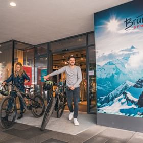 Two cyclists with mountain bikes in front of a Bründl sports shop with a large advertising poster of a snow-covered mountain landscape.