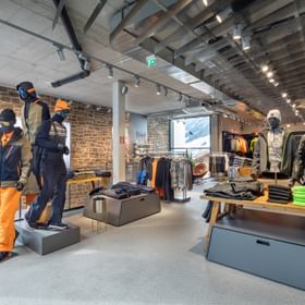 Das Bild zeigt einen Verkaufsbereich in einem Bründl Sports Shop, in dem Schaufensterpuppen in Skibekleidung präsentiert werden. Die Kleidung ist farbenfroh, mit Akzenten in Orange und Grün. Im Hintergrund sind weitere Winterbekleidungsartikel auf Tischen und Kleiderständern ausgestellt, die Wände bestehen aus dekorativem Backstein.