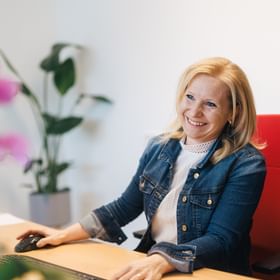 A woman sits on a red office chair at a desk holding a computer mouse. She is smiling, with a keyboard in front of her and a plant visible in the background.