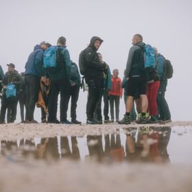 Das Bild zeigt eine Gruppe von Wanderern, die in Outdoor-Kleidung und mit Rucksäcken auf einem nebligen Berggipfel stehen und sich unterhalten.