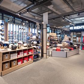 Zu sehen ist ein Verkaufs- und Verleihbereich im Shop Bründl Sports Penkenbahn Talstation Mayrhofen mit vielen Skihelmen, Skibrillen, gefalteter Kleidung und Ski an der rechten Wand. Im Hintergrund sind die Texte „RENT“ und „HEAD“ sichtbar, außerdem zeigt ein Bildschirm eine Wintersportaufnahme.