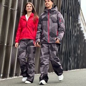 Zwei Mitglieder des österreichischen Olympiateams präsentieren die neue AlphaTauri Teamkleidung für Olympia 2026 in Rot und Grau vor moderner Architektur.