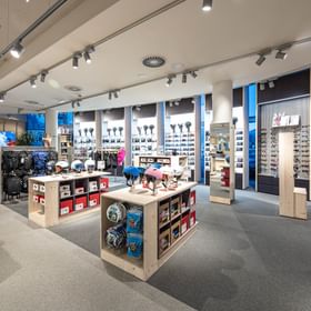 The image shows a bright retail area inside the Bründl Sports AreitXpress valley station shop with ski helmets, ski goggles, backpacks, and other winter sports accessories displayed on shelves, walls, and wooden units. Several helmets are presented on a display table in the foreground, with more helmet and goggle shelves and large windows visible in the background.