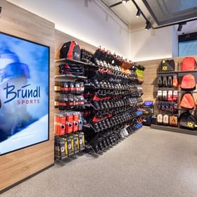 Zu sehen ist ein Skischuhbereich im Shop Bründl Sports Penkenbahn Talstation Mayrhofen mit vielen Skischuhen an Wandregalen sowie Wintersport-Zubehör, Taschen und Rucksäcken. Links ist ein Bildschirm mit dem „Bründl Sports“ Logo.