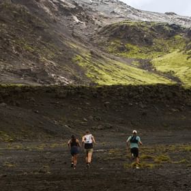 Drei Läufer rennen durch eine weite vulkanische Berglandschaft mit steilen Hängen und moosbewachsenen Felsen.