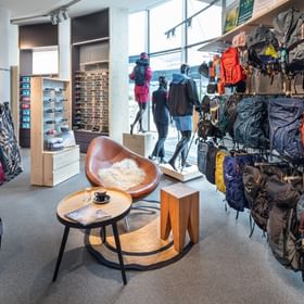 The image shows a retail area inside the Bründl Sports AreitXpress valley station shop with many backpacks displayed on the right wall and outdoor clothing, shorts, and shoes on the left. In the center, mannequins wear hiking clothing, with a small seating area including a chair, table, and stool in front.