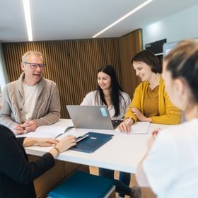 People of the HR team are standing at a table in a modern room, talking to each other. A laptop is placed in the center, with documents and a notebook nearby, while some of the people are smiling and writing.