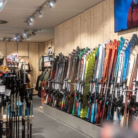 The image shows a retail area inside the Bründl Sports AreitXpress valley station shop with many skis in different colors displayed along a wooden wall. Ski poles and other winter sports accessories are visible in the foreground, with ski clothing and jackets on the left and in the background; a screen above the ski wall shows a winter sports scene.