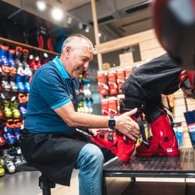Ein Bründl Sports Mitarbeiter hilft einem Kunden beim Anpassen von Skischuhen. Der Mitarbeiter sitzt vor einer Wand mit bunten Skischuhen und sorgt für die perfekte Passform am Fuß des Kunden.