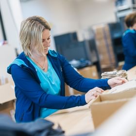 A woman stands at a worktable placing folded clothing into a box. In the background, more clothing items and another person working can be seen.