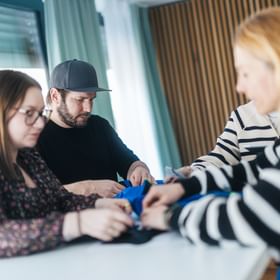 Four people stand at a table holding a blue piece of clothing together. They look at it with focus while examining or working on it.