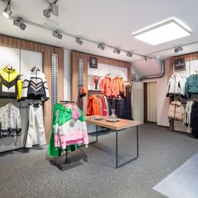 Zu sehen ist ein Verkaufsbereich im Shop Bründl Sports Saalbach life.style mit hochwertiger Skibekleidung, Winterjacken, Hosen und Accessoires an Wänden, Ständern und einem Tisch. Im Bild sind die Marken- und Schriftzüge „Bogner“ und „Moncler“ sichtbar; rechts ist ein Teil einer Rolltreppe zu sehen.