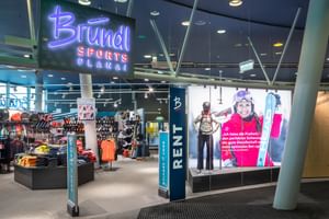 Das Bild zeigt den Eingang eines Bründl Sports Shops in der Planai mit einem großen, leuchtenden Bründl Sports Schild. Im Inneren sind verschiedene Wintersportprodukte wie Kleidung und Ausrüstung ausgestellt, und es gibt einen Bereich für Verleih und Service. Ein großes Werbeposter zeigt eine lächelnde Skifahrerin.