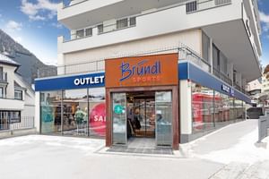 Zu sehen ist der Eingang eines Sportgeschäfts mit dem Schriftzug „Bründl Sports“ über der Tür. In den großen Schaufenstern stehen Schaufensterpuppen und es sind gut sichtbar die Wörter „OUTLET“ und „SALE“ zu lesen.