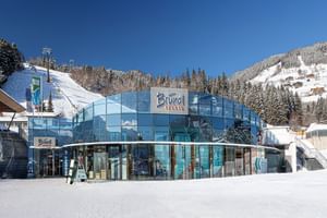 The image shows the Bründl Sports AreitXpress valley station shop with a large blue glass facade next to a snowy ski slope. The “Bründl Sports” logo and the texts “SKIDEPOT,” “SHOP,” “RENT,” “DEPOT,” and “SERVICE” are visible on the building, with a ski lift, forest, and snow-covered mountains in the background.