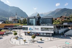 The Kitzsteinhorn Kaprun Center stands in front of an alpine mountain backdrop. A Bründl Sports shop is located inside the building, with signs reading “KAPRUN CENTER,” “Bründl Sports,” “Kitzsteinhorn,” “Tickets,” and “Ski Depot.”