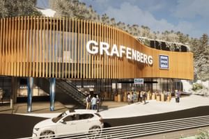 A modern building with a curved wooden facade and the text “GRAFENBERG” stands in front of snow-covered trees. Several people stand near the entrance, a car is passing by, and signs reading “Flachau Snow Space Salzburg,” “Bründl Sports,” and “Ticketschalter” are visible, while a cable car runs above.