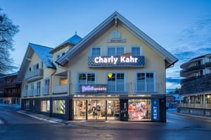 Das Bild zeigt ein Gebäude mit dem "Bründl Sports" Shop im Erdgeschoss und einem Schild für "Charly Kahr" im oberen Bereich, umgeben von anderen Gebäuden in einer winterlichen Umgebung.
