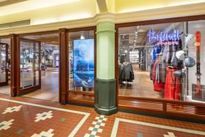 The image shows the glass storefront of the Bründl Sports McArthurGlen Designer Outlet Salzburg shop with a visible “Bründl Sports” logo in the window. Sports clothing, skis, helmets, and accessories are visible inside; a screen shows the text “#BRÜNDLFAN,” and “kassa” can also be seen in the background.