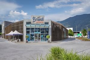 Außenansicht der Bründl Sports Filiale in Saalfelden. Vor dem modernen Geschäftsgebäude mit großem Bründl Sports Logo stehen mehrere Fahrräder unter zwei weißen Zelten. Im Hintergrund sind Berge, Bäume und ein teilweise bewölkter Himmel zu sehen. Sichtbarer Text: „Bründl Sports“ und „Einfahrt“.