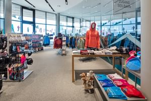 Das Bild zeigt eine Verkaufsfläche eines Bründl Sports Shops mit einer Auswahl an Wintersportbekleidung, Accessoires und Souvenirs. Im Vordergrund steht eine Puppe mit einem Hoodie, daneben sind T-Shirts und weitere Kleidungsstücke ausgestellt. Große Fenster bieten einen Blick nach draußen auf die winterliche Landschaft.