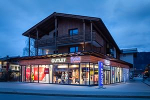 Das Bild zeigt das Bründl Sports Outlet bei Dämmerung. Das Gebäude ist beleuchtet, was die "Outlet" und "Bründl Sports" Schilder gut sichtbar macht. Das Schaufenster zeigt sportliche Winterbekleidung.