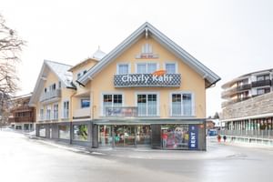Das Bild zeigt ein Gebäude mit dem "Bründl Sports" Shop im Erdgeschoss und einem Schild für "Charly Kahr" im oberen Bereich, umgeben von anderen Gebäuden in einer winterlichen Umgebung.