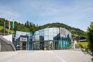 Zu sehen ist der Bründl Sports AreitXpress Talstation Shop mit großer Glasfassade, Shop-Eingang und sichtbarem „Bründl Sports“ Schriftzug an der Fassade. Links befinden sich Fahnen und eine Treppe, im Hintergrund sind Wald, Wiesen und ein Berg zu sehen; am Gebäude steht außerdem der Text „SKIDEPOT“.