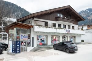 Das Bild zeigt einen Bründl Sports Shop in einem alpenländischen Gebäude mit Schaufenstern, einem Auto davor und Bergen im Hintergrund.