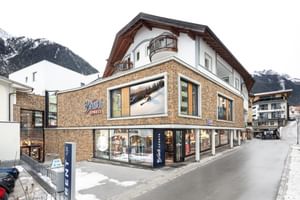 Bründl Sports Ischgl Zentrum in daylight, featuring a stone façade, large display windows and Bründl Sports logos. A “RENT” sign stands outside the store, with patches of snow along the street and snow-covered mountains in the background.