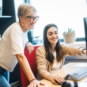 Eine Frau sitzt vor einem Computer und zeigt auf den Bildschirm, während eine andere Frau daneben steht und lächelt. Auf dem Schreibtisch stehen Monitor, Tastatur und Maus, im Hintergrund sind Ordner und eine Pflanze zu sehen.