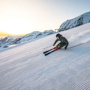 Das Bild zeigt einen Skifahrer, der bei Sonnenaufgang eine schneebedeckte Piste hinunterfährt, umgeben von Berglandschaft.