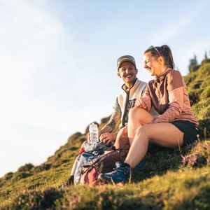 Two people are sitting on a grassy slope, laughing together. Backpacks and a water bottle lie beside them, with hills and trees visible in the warm sunlight in the background.