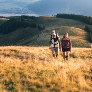 Two people with backpacks walk uphill through tall, golden grass. In the background, forested hills, a lake, and a valley are visible in the warm light of the sunset.