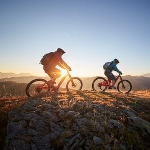 two mountainbikers are riding on a mountain ridge, while the sun sets in the background