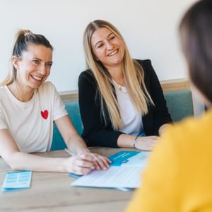 Zwei Frauen sitzen nebeneinander an einem Tisch und lächeln einer dritten Person gegenüber, die im Vordergrund unscharf zu sehen ist. Auf dem Tisch liegen ein Dokument und ein Stift, den eine der Frauen in der Hand hält.