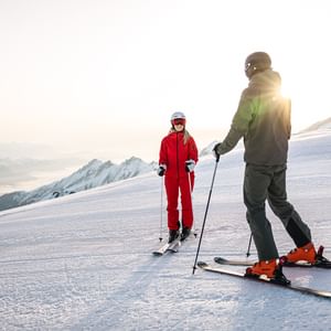 Das Bild zeigt zwei Skifahrer auf einer verschneiten Piste, umgeben von Bergen. Eine Person trägt eine rote Skiausrüstung, die andere eine grüne. Sie unterhalten sich bei Sonnenaufgang oder Sonnenuntergang in einer idyllischen Winterlandschaft.