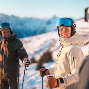 Drei Skifahrer stehen mit Helm und Skibrille auf einer verschneiten Piste und lachen gemeinsam. Im Hintergrund sind schneebedeckte Berge im warmen Morgenlicht zu sehen.
