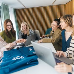 Mehrere Frauen stehen und sitzen an einem Tisch mit geöffneten Laptops und betrachten blaue Kleidungsstücke. Eine Frau zeigt auf ein Detail, während die anderen lächeln und auf die Kleidung schauen.