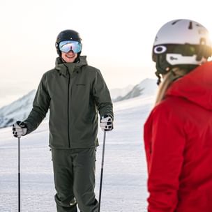 Das Bild zeigt zwei Personen auf einer Skipiste. Beide tragen Skibekleidung und Helme, eine Person in Grün, die andere in Rot. Sie scheinen eine entspannte Unterhaltung in der verschneiten Berglandschaft zu führen.