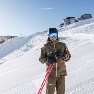 Ein lächelnder Skifahrer in brauner Skibekleidung, schwarzem Helm und blau verspiegelter Skibrille hält rote Skistöcke. Im Hintergrund sind eine präparierte Skipiste, verschneite Hänge und Berggebäude unter blauem Himmel zu sehen.