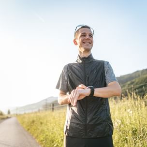Läufer steht auf einem asphaltierten Weg in alpiner Landschaft und blickt lächelnd zur Seite, während er auf seine Sportuhr schaut.