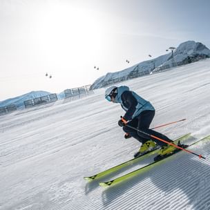 Ein Skifahrer in blauer Skiausrüstung fährt eine frisch präparierte Piste hinunter. Im Hintergrund sind schneebedeckte Berge, ein Seilbahnträger und Gondeln zu sehen.